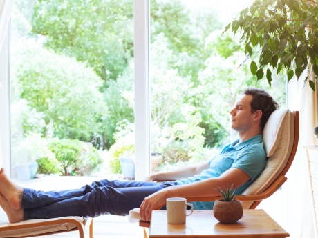 Early 30s man reclining in chair inside home with glass doors looking out into the yard behind him
