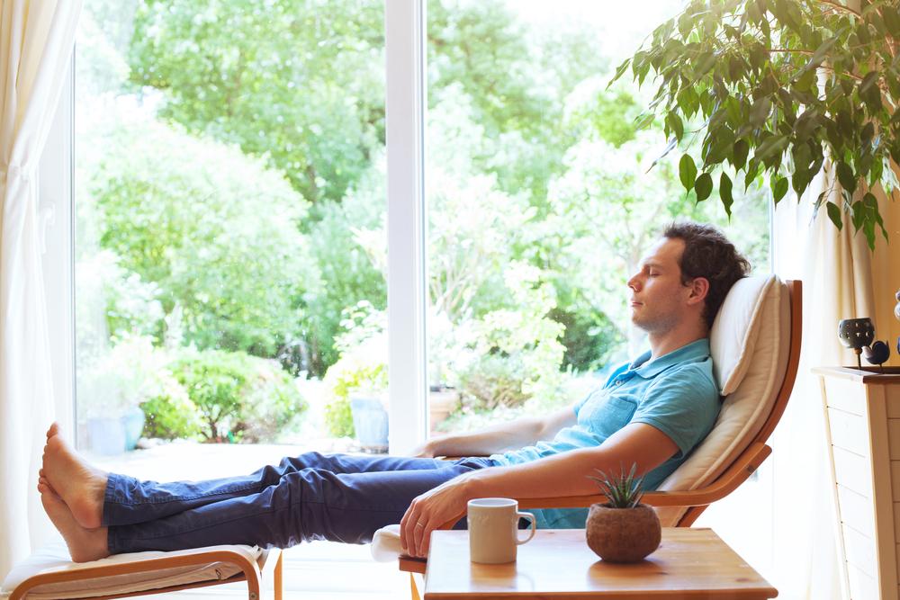 Early 30s man reclining in chair inside home with glass doors looking out into the yard behind him